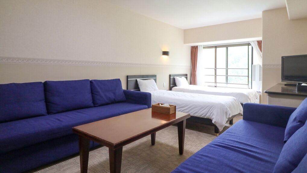 Une chambre d’hôtel avec un canapé bleu, une table en bois, deux lits, une télévision et une fenêtre avec des rideaux.