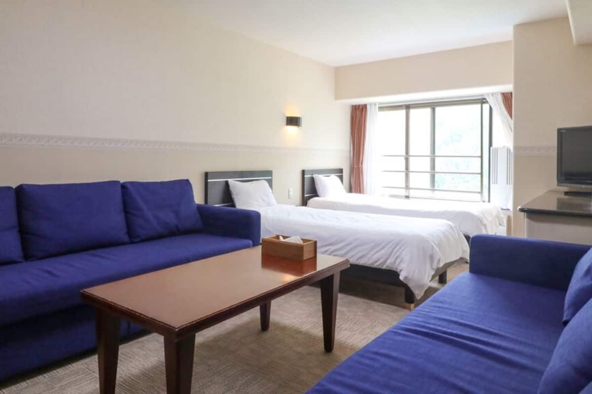 A hotel room with a blue sofa, a wooden table, two beds, a television, and a window with curtains.