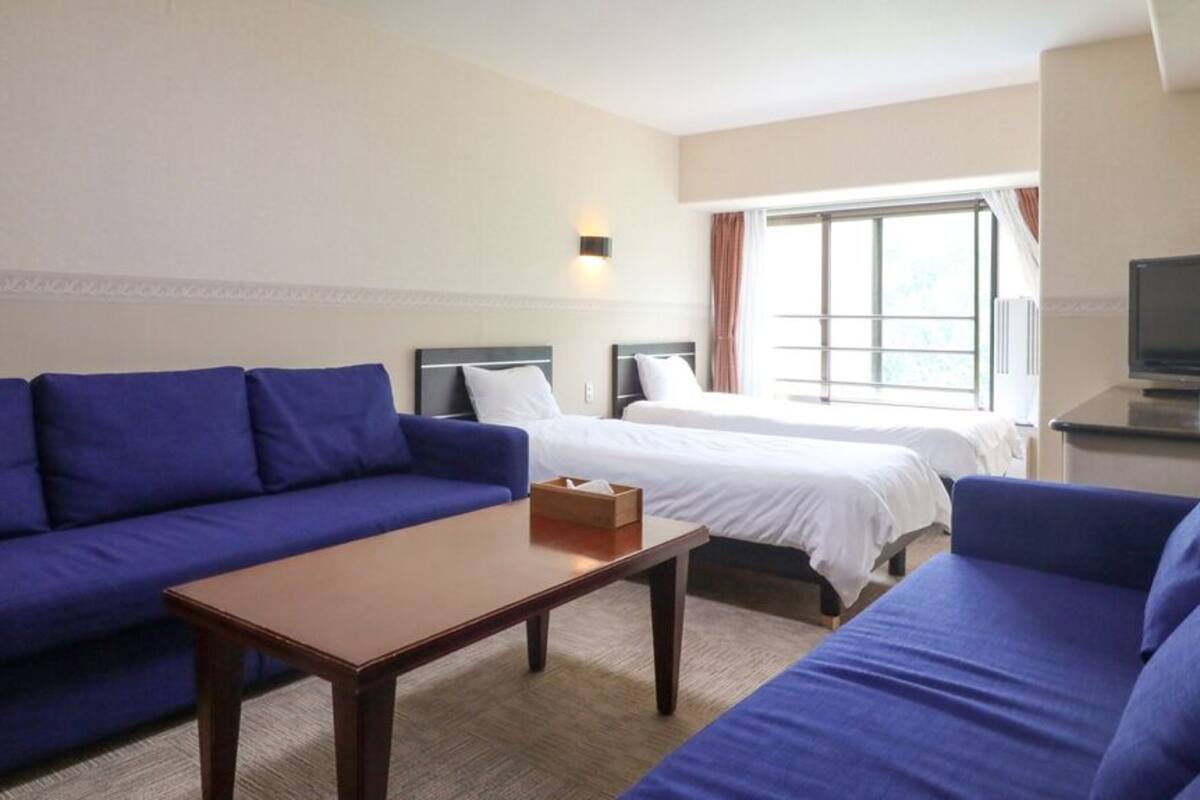Une chambre d’hôtel avec un canapé bleu, une table en bois, deux lits, une télévision et une fenêtre avec des rideaux.