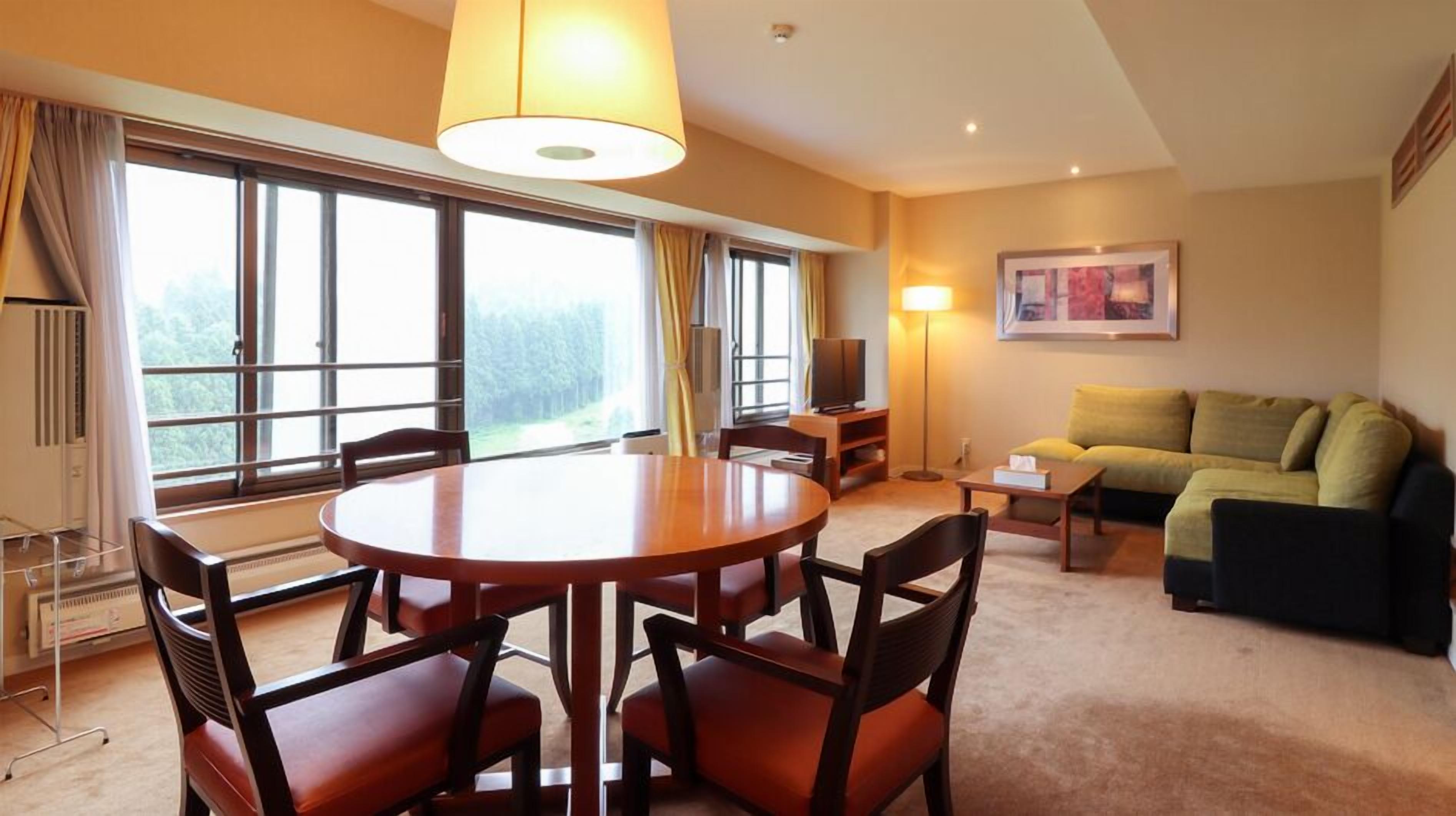 A hotel room with a round dining table, four chairs, a sofa, a TV, and large windows with curtains.