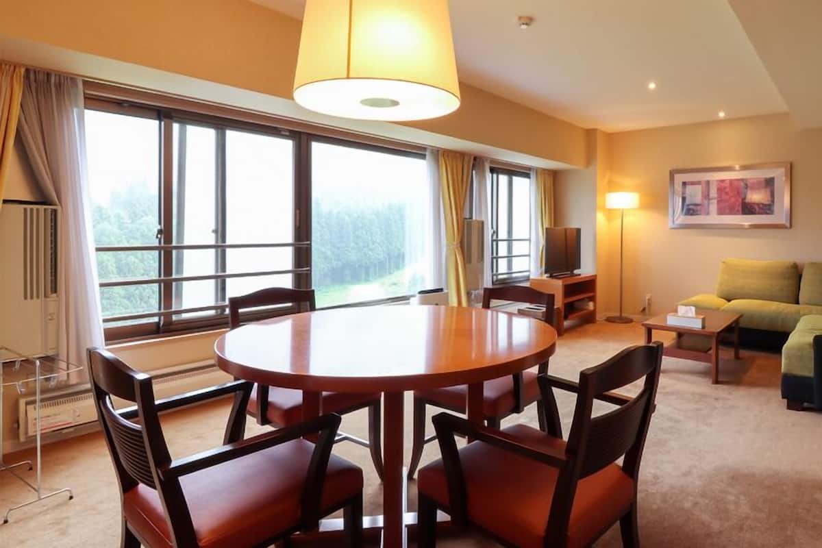 A hotel room with a round dining table, four chairs, a sofa, a TV, and large windows with curtains.