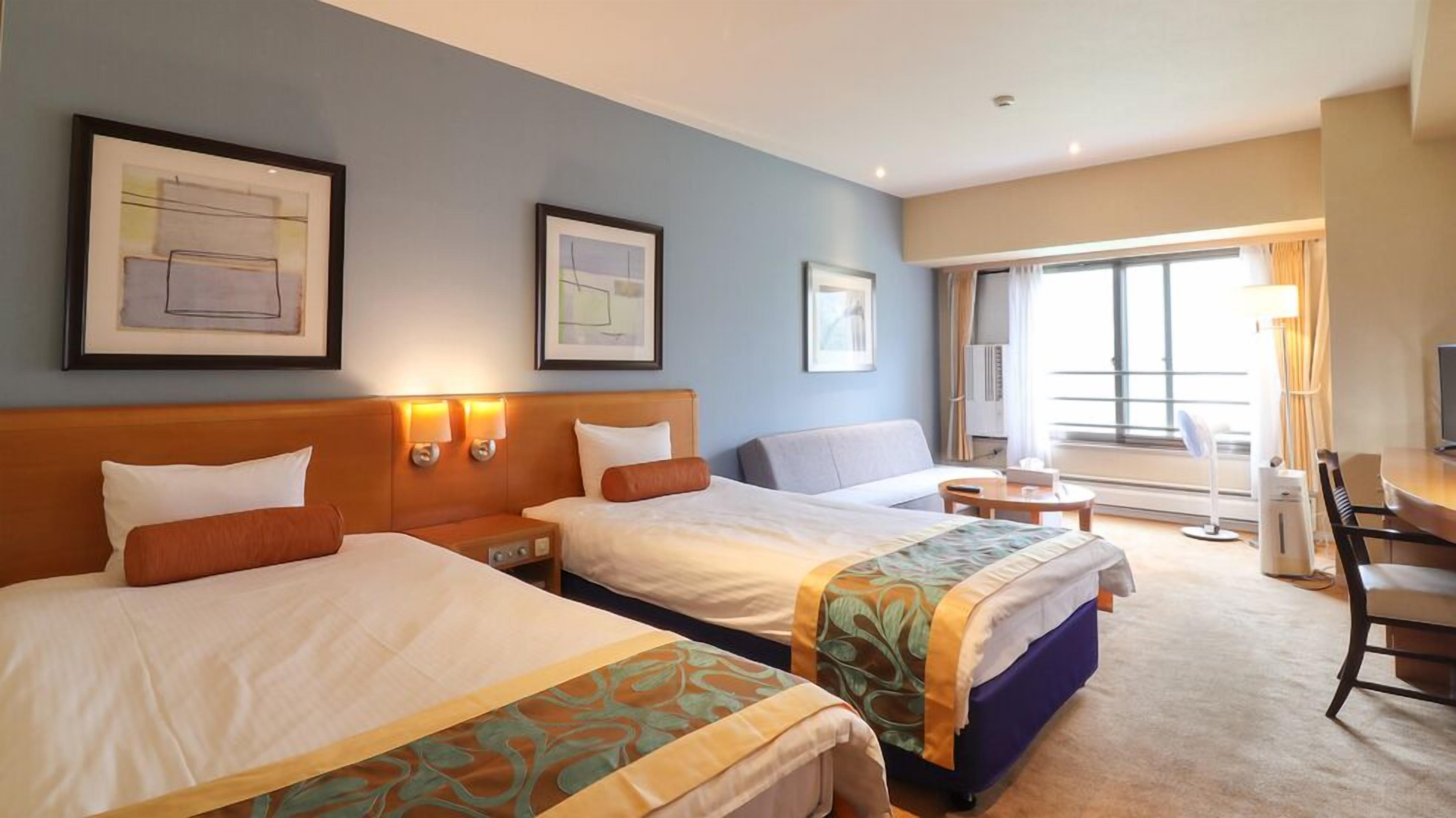 Une chambre d’hôtel avec deux lits, un bureau, une chaise et une grande fenêtre.