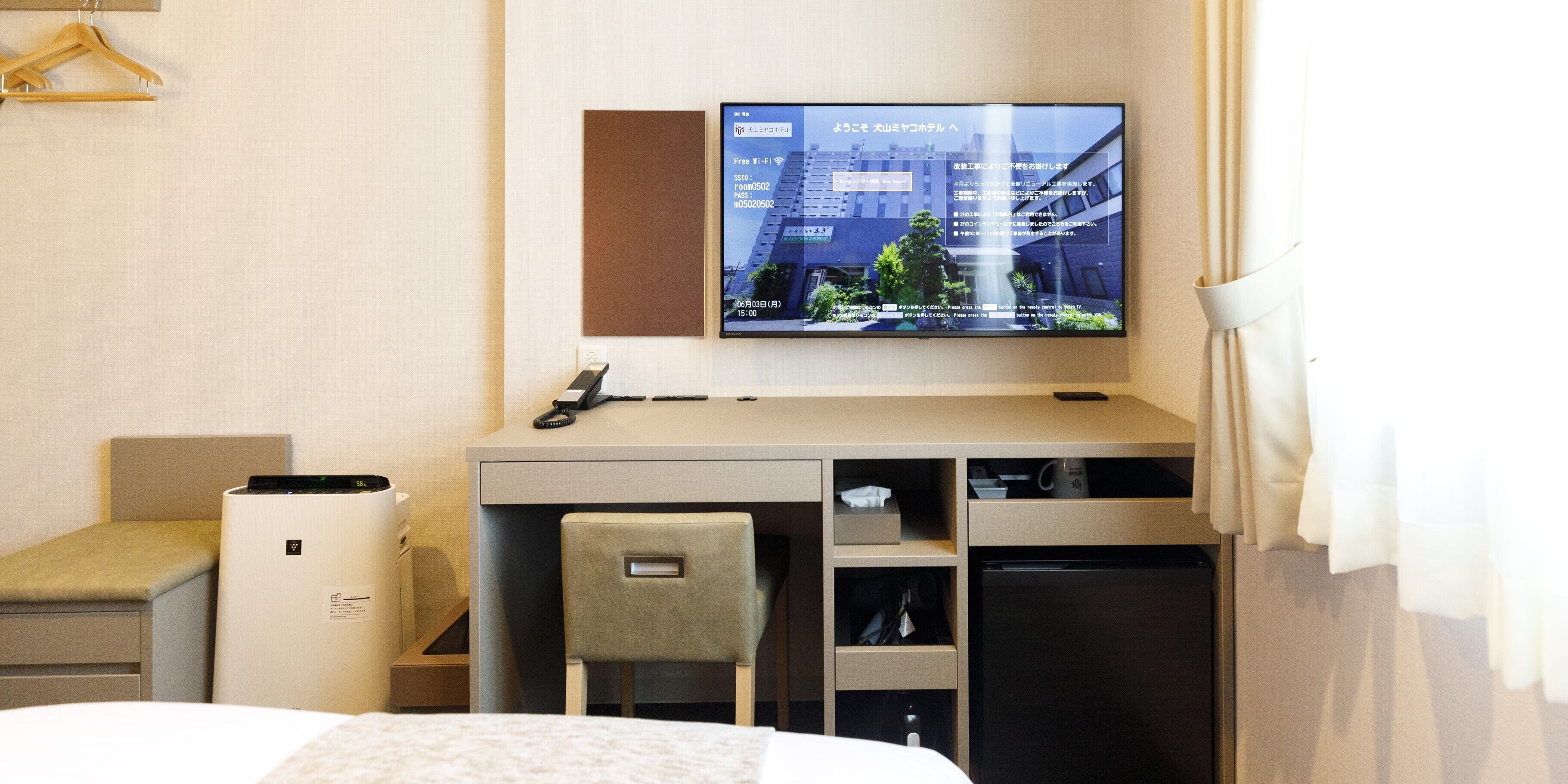 A hotel room with a bed, a desk with a chair, a television mounted on the wall, and a small refrigerator.