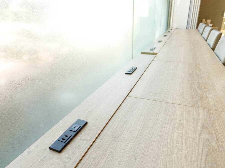 A conference table with a power strip and a remote control, set against a glass wall with natural light.