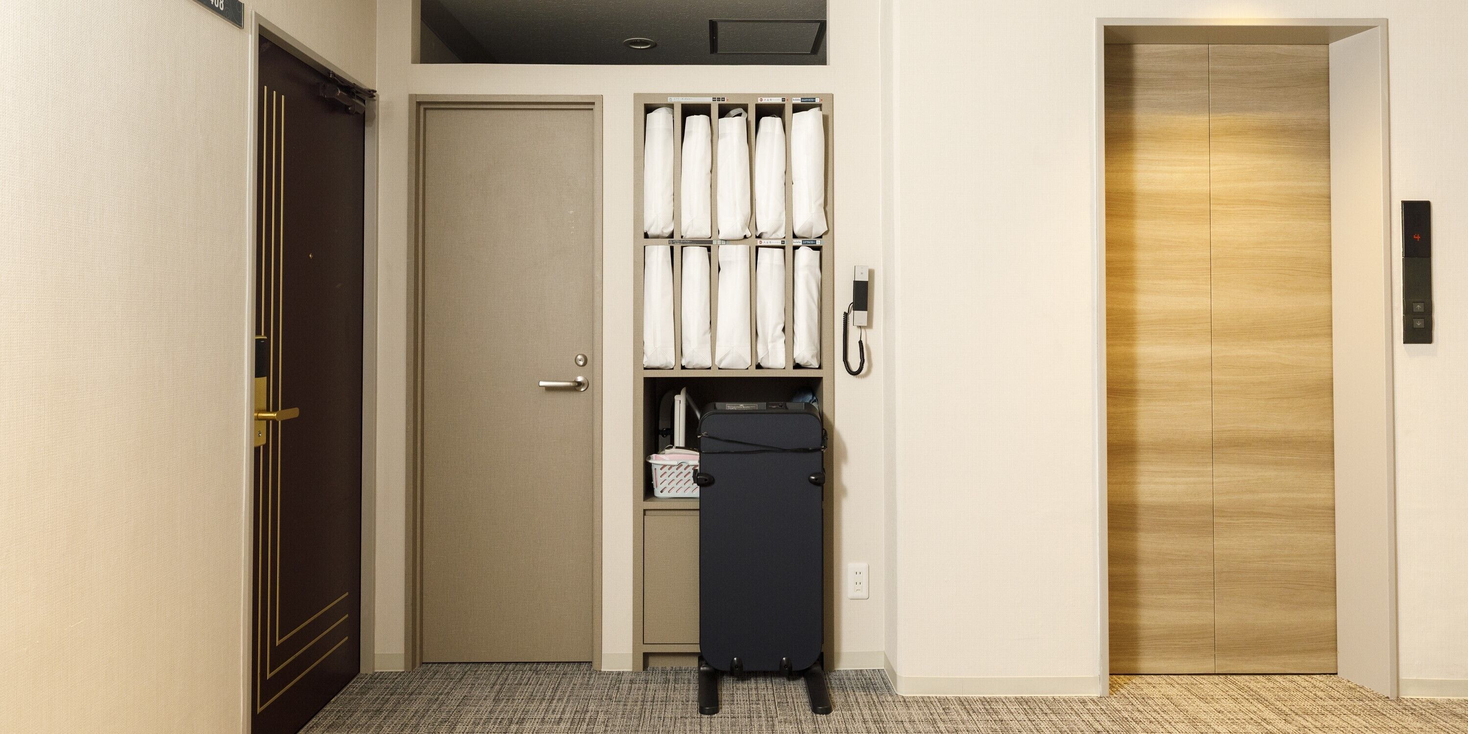 A hotel room with a closed door, a wardrobe with hanging robes, a suitcase, and a wall-mounted phone.