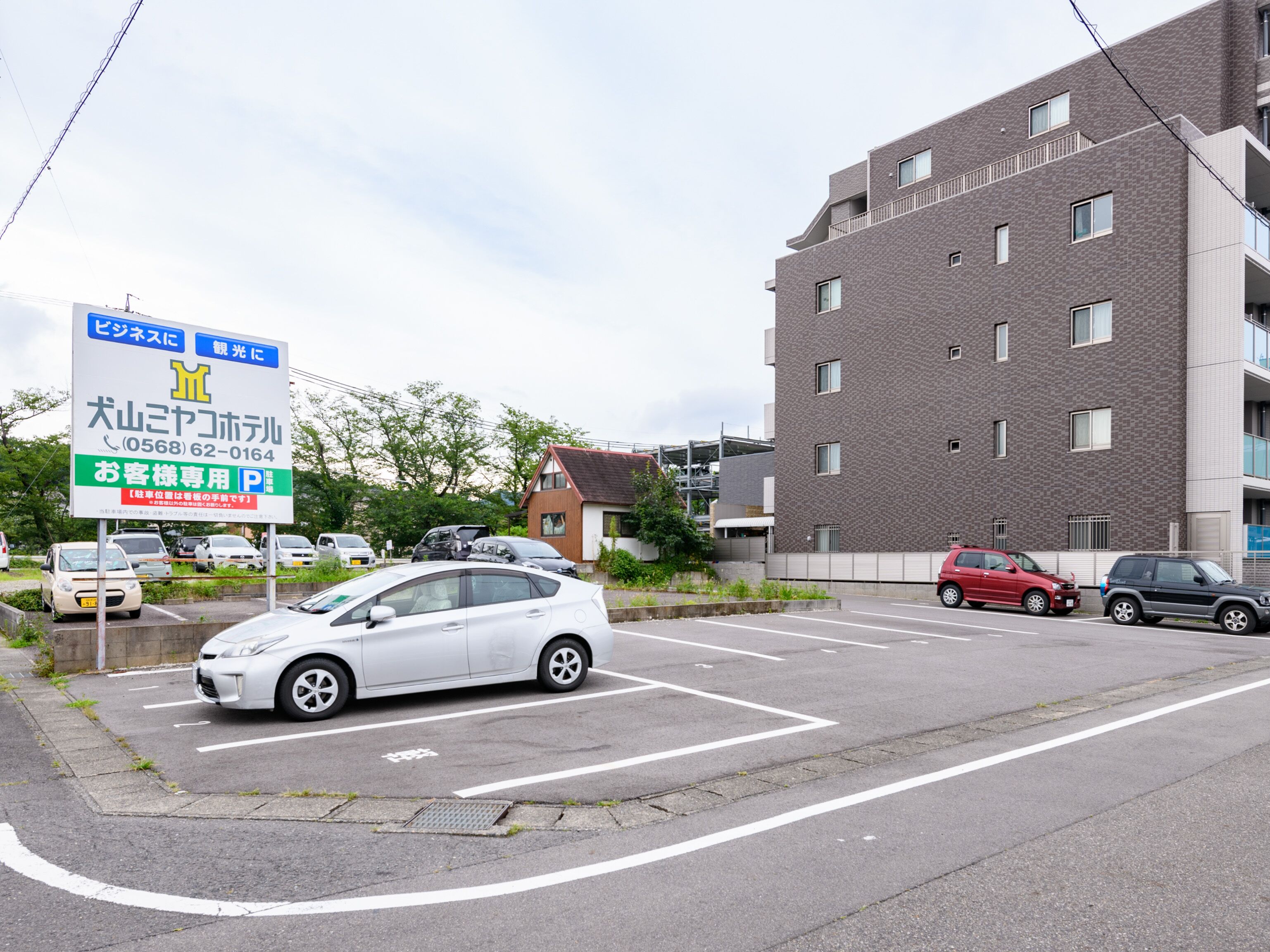 A parking lot with several cars, a building with multiple windows, and a signboard with Japanese text.