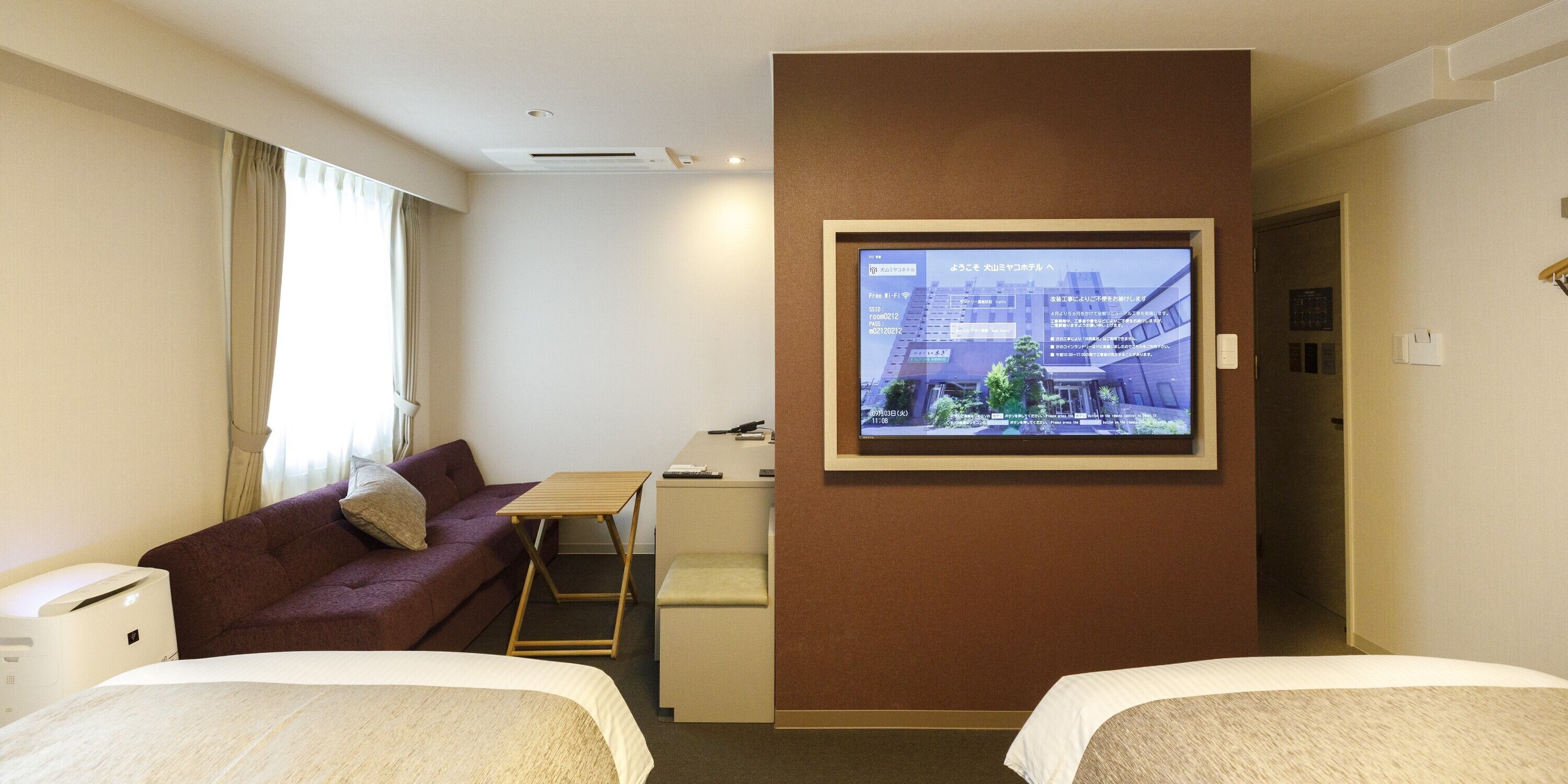 A hotel room with two beds, a desk, a TV mounted on the wall, a sofa, and a window with curtains.