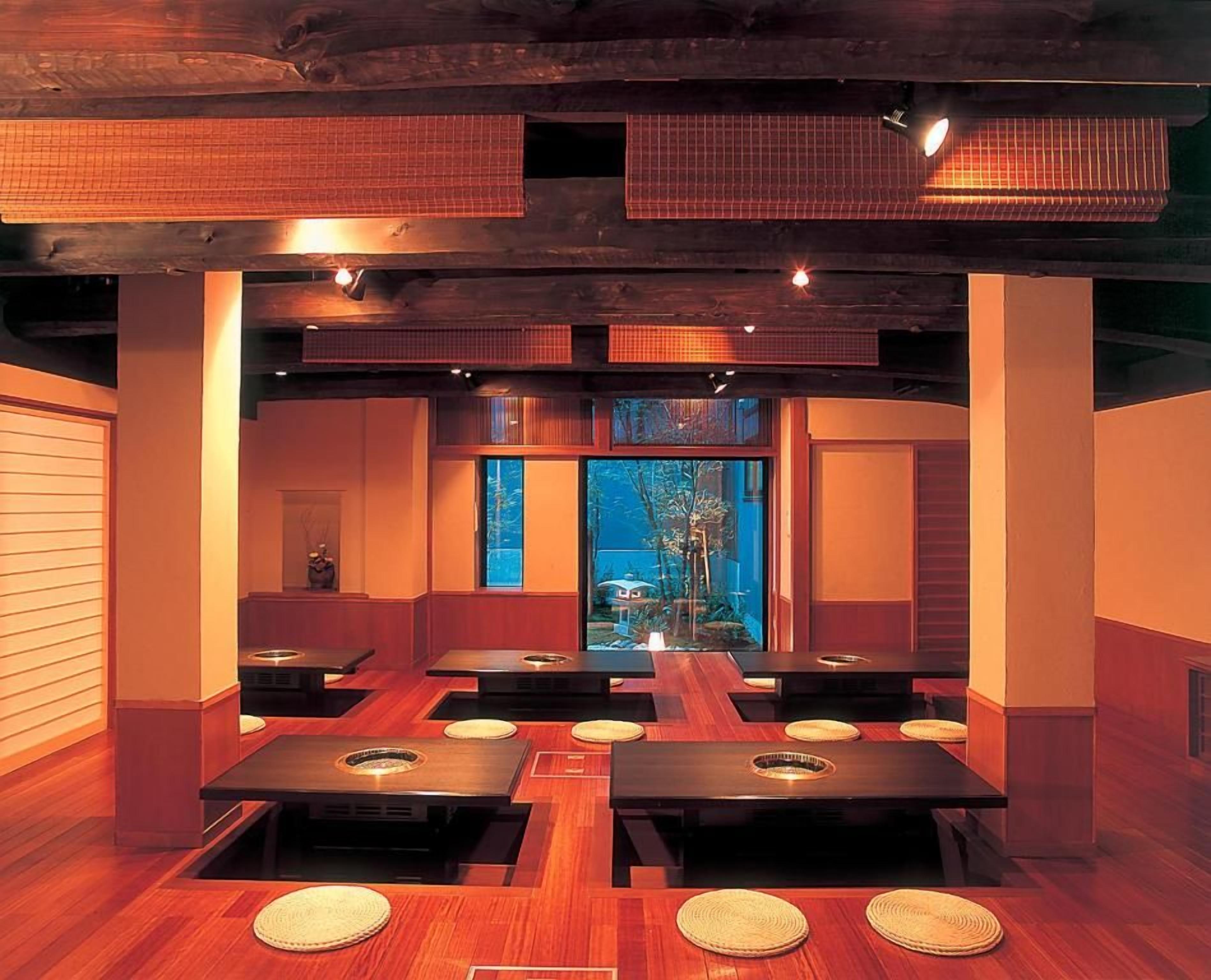 Un restaurante con mesas y sillas de madera, vista a árboles a través de las ventanas y techo con vigas a la vista.