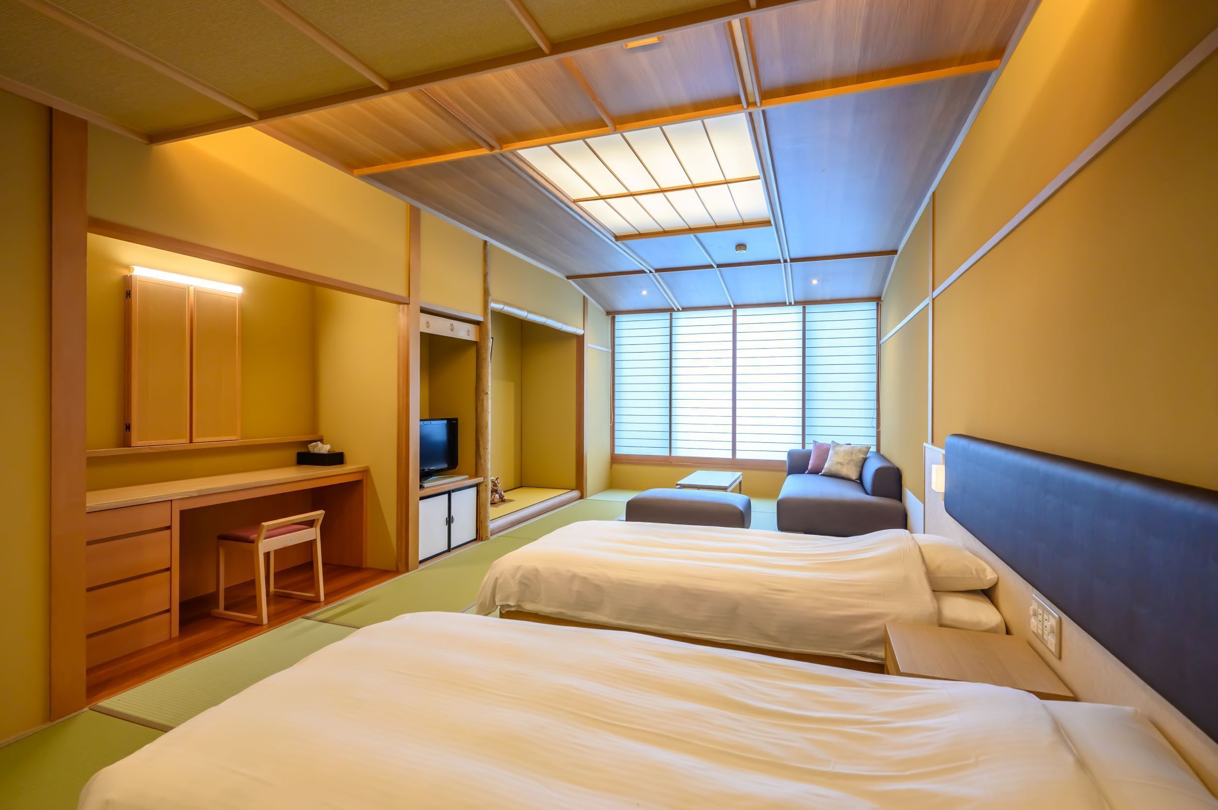 A hotel room with two beds, a desk, a television, and a window with blinds.