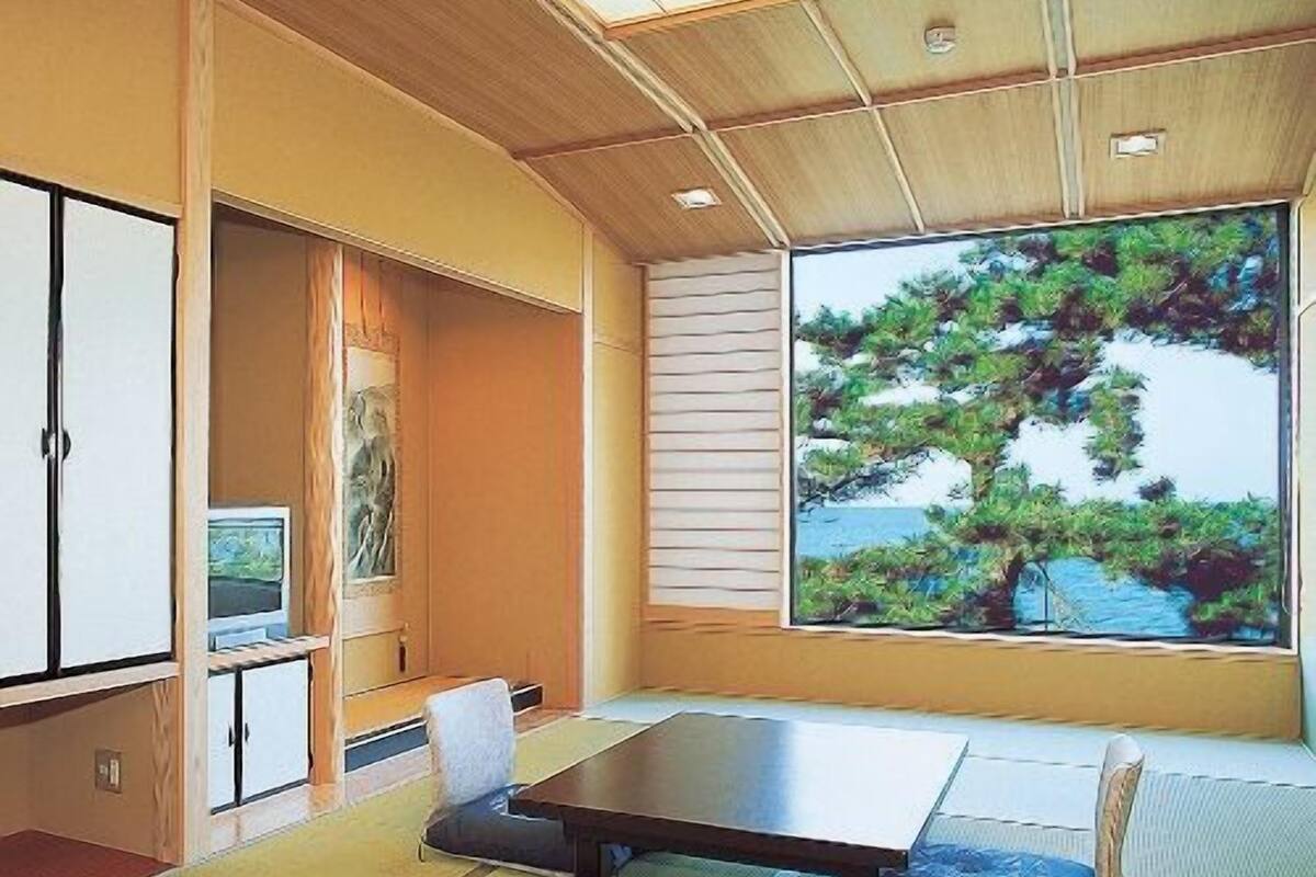 A tatami mat room with a low table, a chair, a cabinet, and a large window with a view of trees.