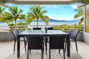 A balcony with a glass table and chairs, overlooking a beach and palm trees.