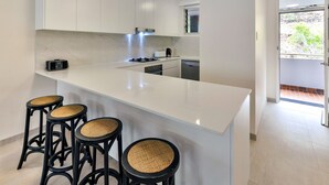 A modern kitchen with white cabinetry, a large white island with bar stools, and a view of the outdoors through a sliding door. - Frangipani 104 (Whitsundays)
