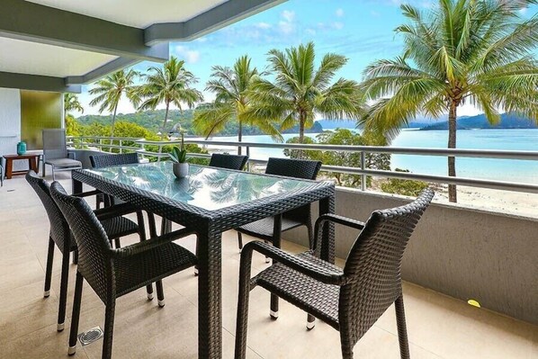 A balcony with a glass table and wicker chairs, overlooking a beach with palm trees and a view of the sea.