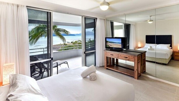 A hotel room with a large bed, a TV, a desk, and a balcony with a view of the ocean. - Frangipani 104 (Whitsundays)