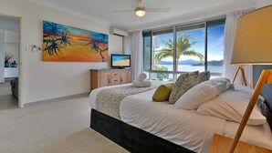 A bedroom with a bed, a television, a painting, and a view of the sea. - Frangipani 104 (Whitsundays)