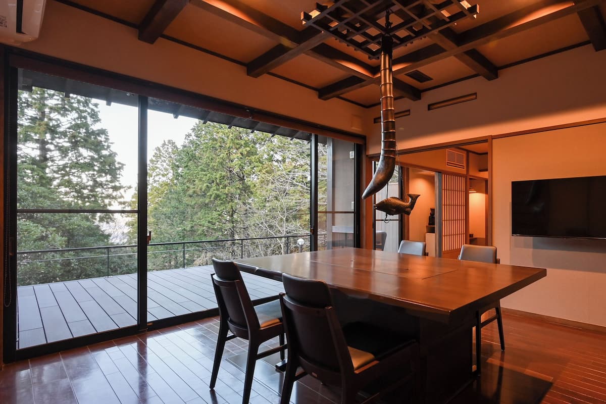 Un comedor moderno con una mesa grande y sillas, un televisor en la pared y vistas al exterior a través de amplios ventanales.