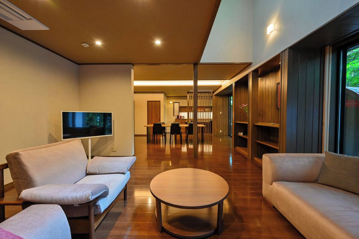 Una sala de estar moderna con un sofá, una mesa de centro y un televisor. Se observa un área de comedor con sillas y una mesa al fondo.