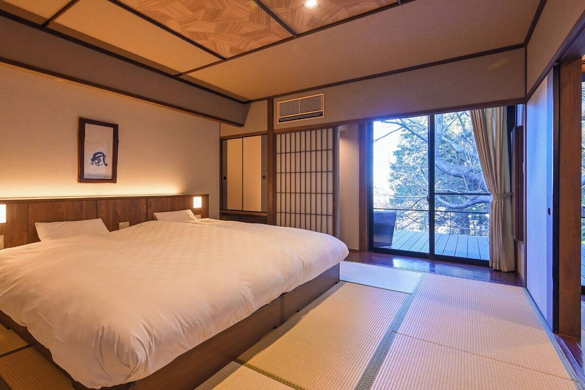 Un dormitorio de estilo japonés tradicional con suelo de tatami, una cama grande, puertas correderas y vistas al exterior.