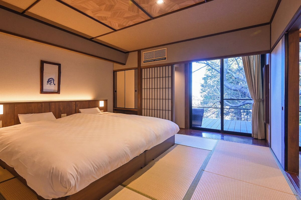 A traditional Japanese-style bedroom with tatami flooring, a large bed, sliding doors, and a view of the outdoors.
