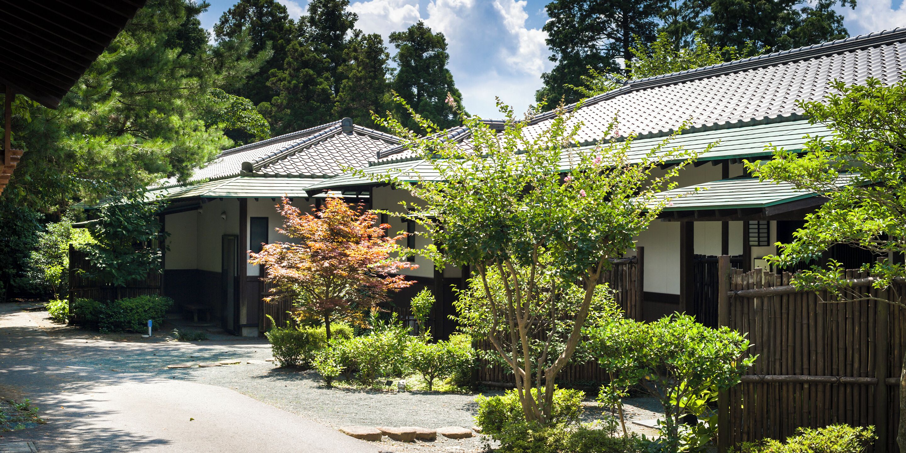 Un edificio de estilo japonés tradicional con techo de tejas y paredes de madera, rodeado de abundante vegetación y un sendero de grava.