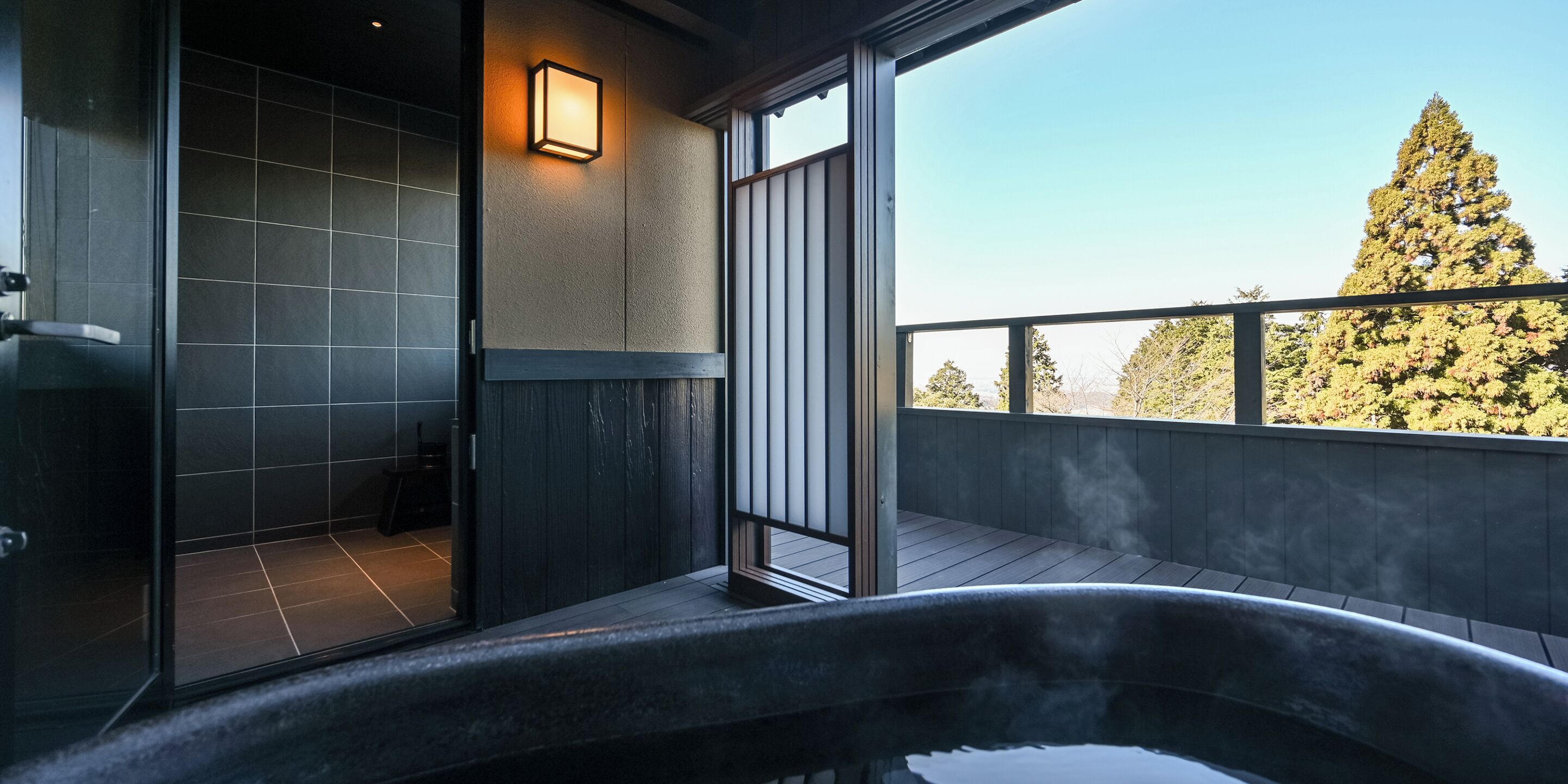 Un baño moderno con ducha, bañera y un amplio ventanal que ofrece vistas a árboles y un edificio.