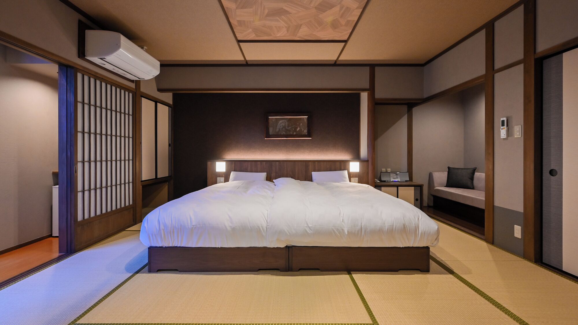 A traditional Japanese-style hotel room with tatami flooring, a large bed, a futon, a shoji screen, and a ceiling fan.
