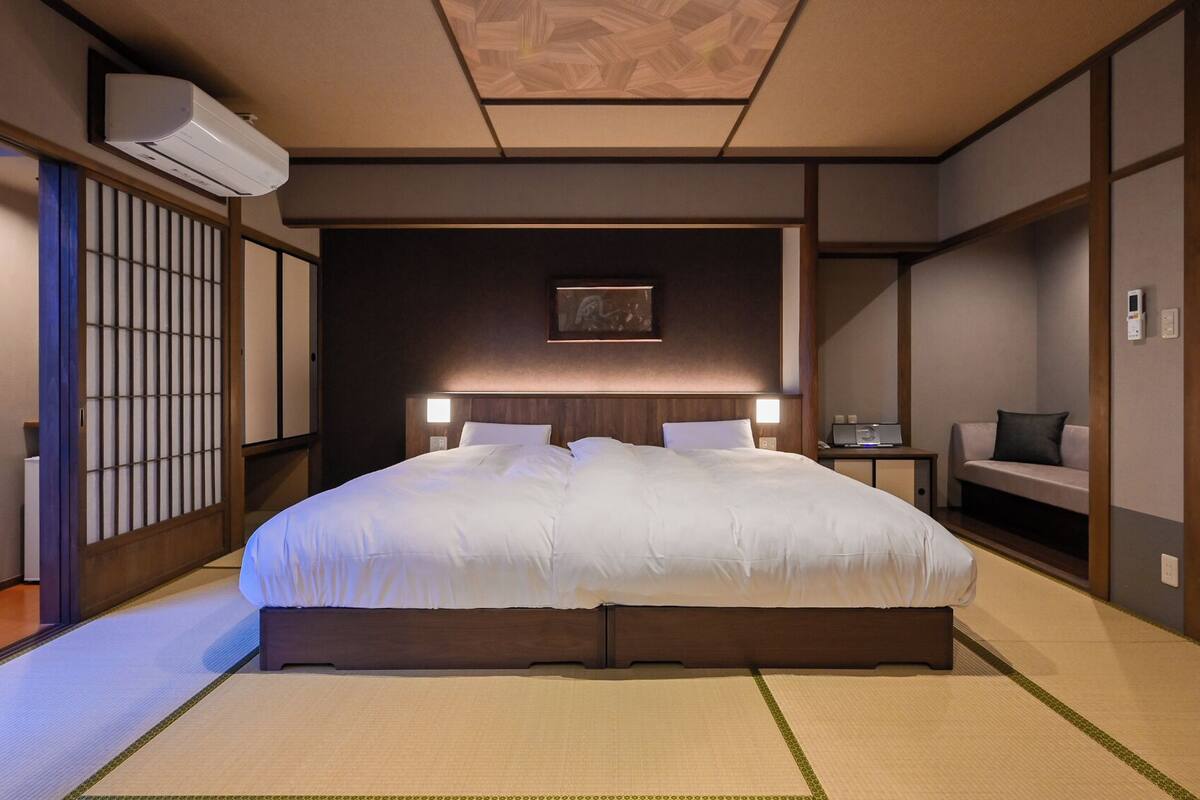 Habitación de hotel tradicional japonesa con suelo de tatami, una cama grande, un futón, un celosía de papel y un ventilador de techo.