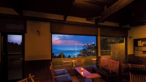 Una habitación acogedora con un ventanal grande que da a un paisaje urbano al atardecer. Hay sillas de madera y una mesita con una vela.
