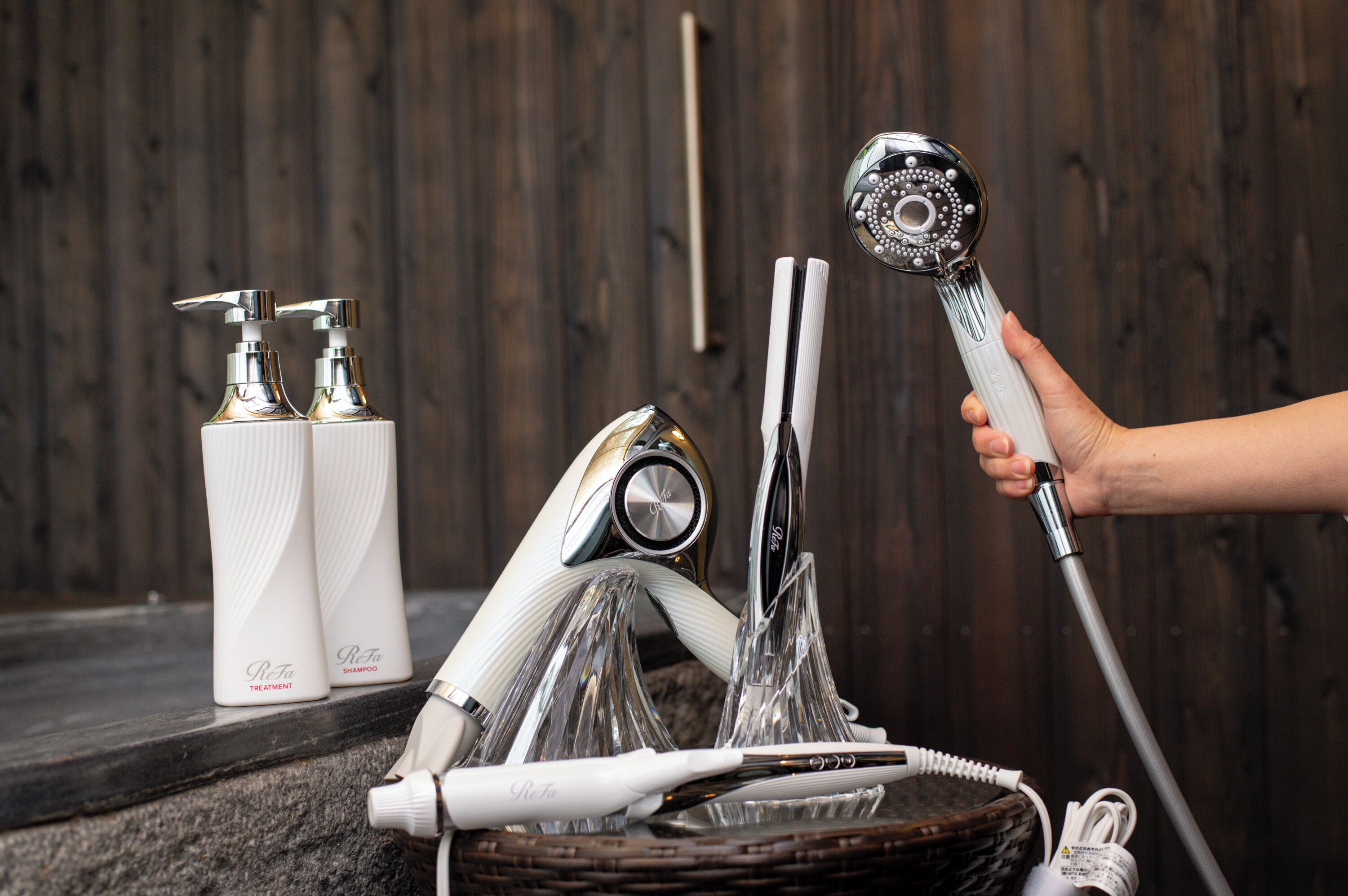 Ein Badezimmer-Waschtisch mit Holzoptik, der eine Hand mit einer Duschbrause, eine Shampooflasche und eine Pflegeflasche zeigt.