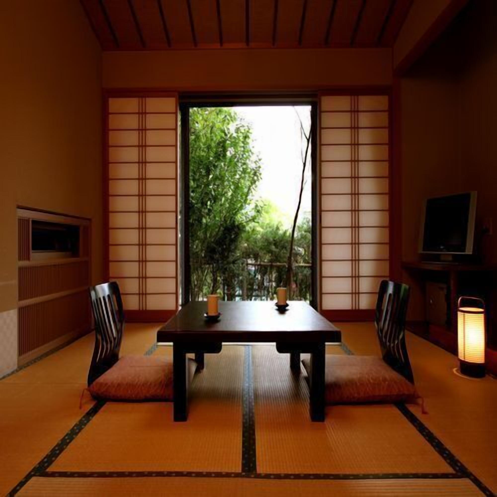 A tatami mat room with a low table, two chairs, a TV, and a window with a view of greenery.