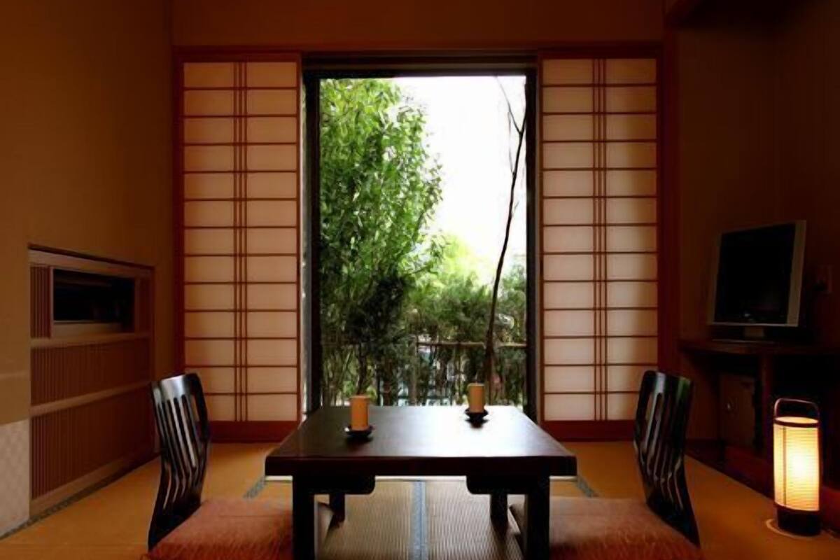 Ein Raum mit Tatami-Matten, einem niedrigen Tisch, zwei Stühlen, einem Fernseher und einem Fenster mit Blick ins Grüne.