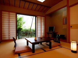 Una habitación japonesa tradicional con suelo de tatami, una mesa baja, sillas, un televisor y una puerta corrediza que da a un balcón.