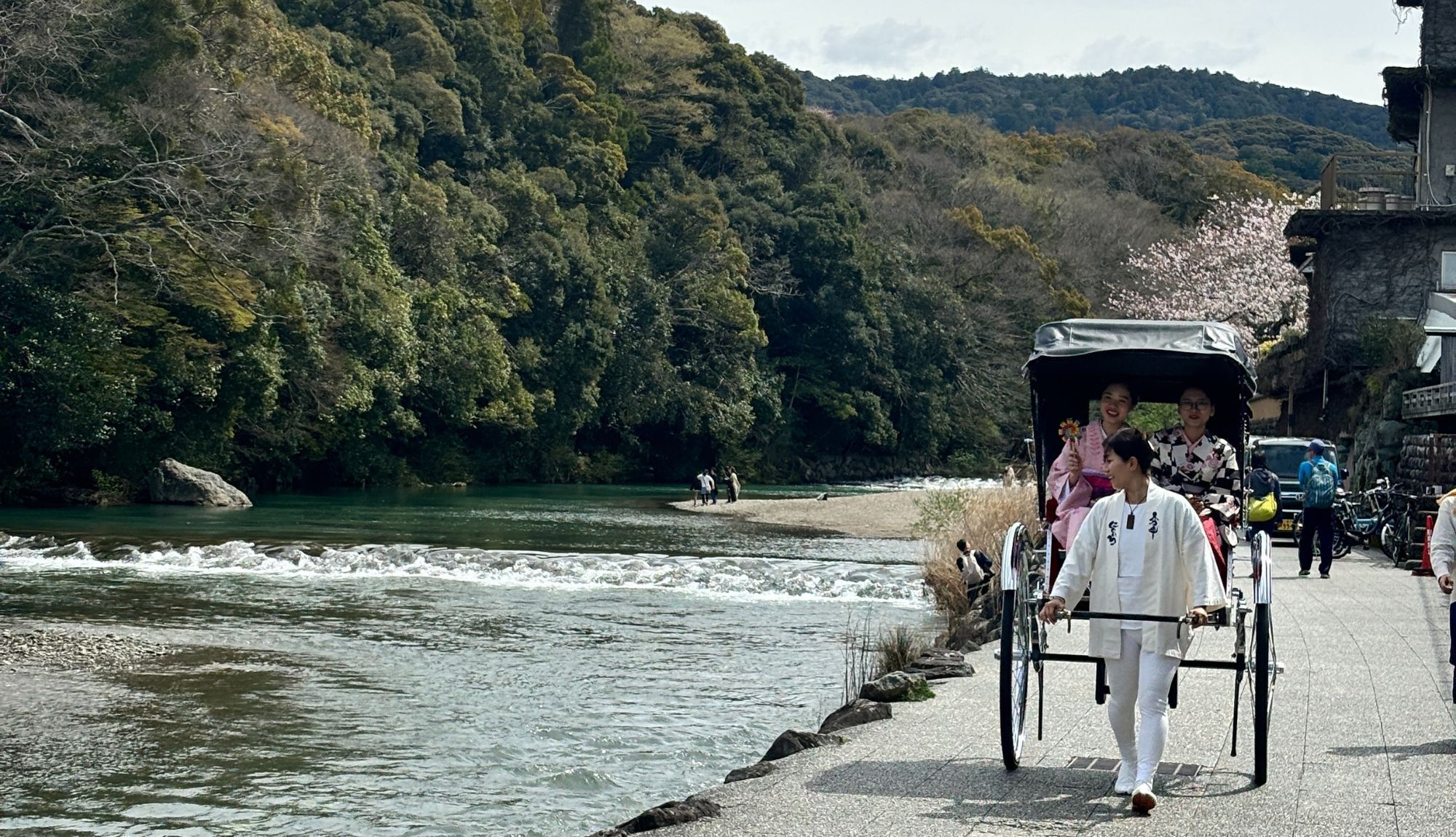 A riverside walkway with a traditional carriage, people walking, and a building with a stone facade.