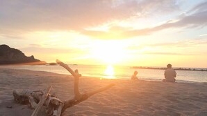 Escena de playa con atardecer, un tronco grande de madera a la orilla y dos personas sentadas en la arena.