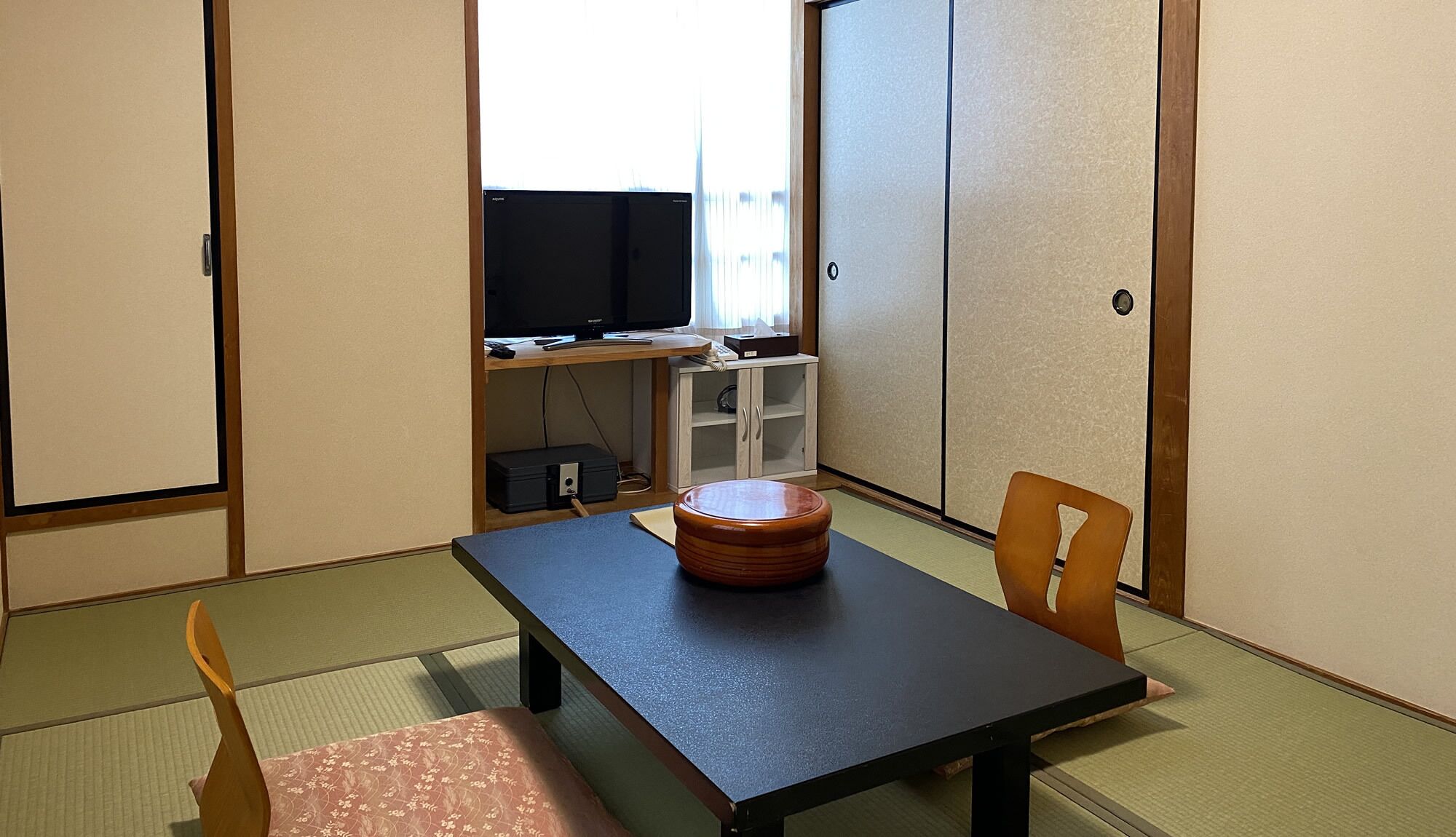 Une pièce de style japonais traditionnel, avec un sol recouvert de tatamis, une petite table basse accompagnée d’un tabouret en bois, un téléviseur posé sur une étagère et des portes coulissantes.