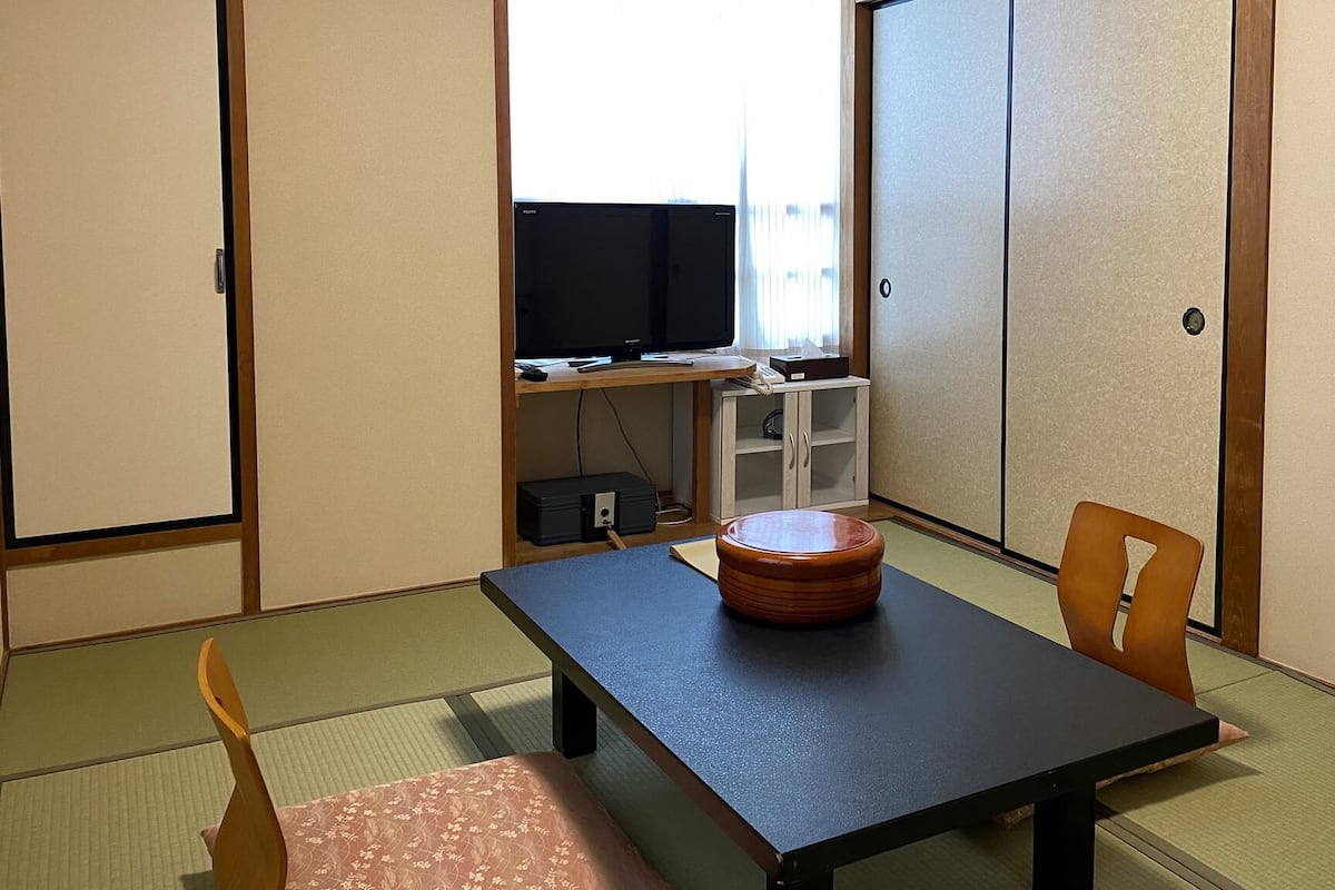 Una habitación japonesa tradicional con suelo de tatami, una mesa baja con un taburete de madera, un televisor en un estante y puertas correderas.