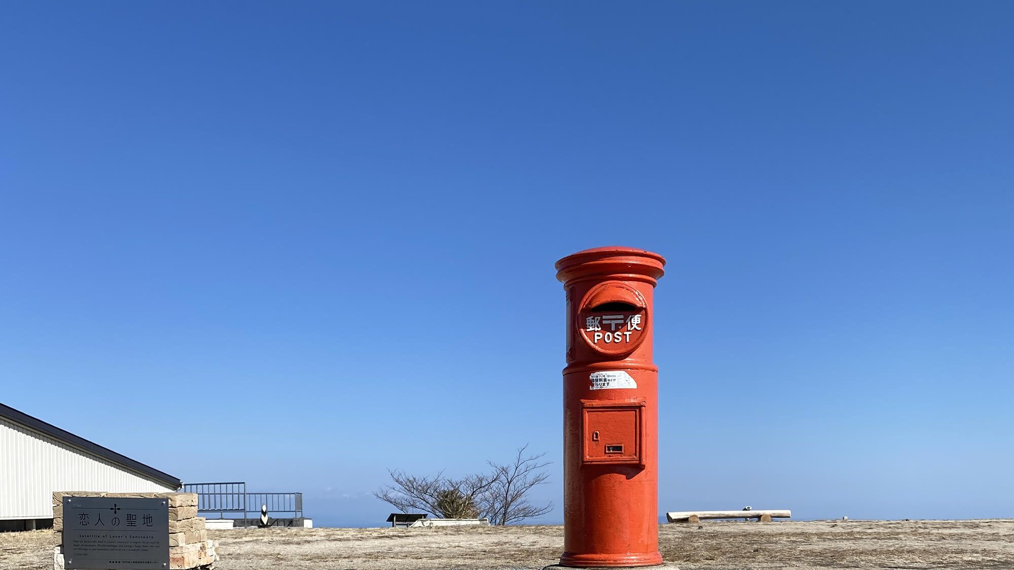 Un palo rosso con le scritte 'POST' e 'POST', una panchina e un edificio sullo sfondo.
