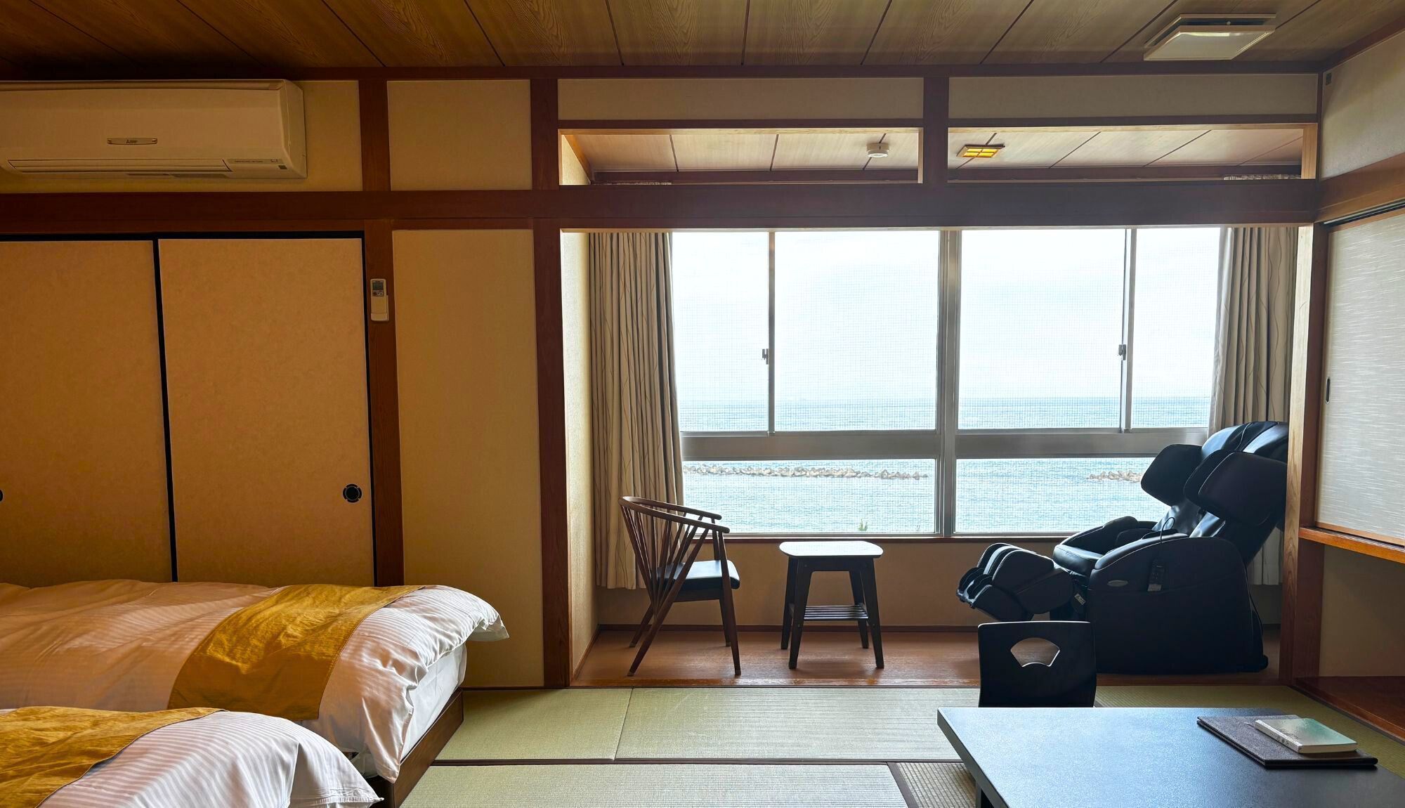 Une chambre d’hôtel comprenant un lit, une chaise, un fauteuil de massage, une petite table et une vue sur la mer grâce à de grandes fenêtres.