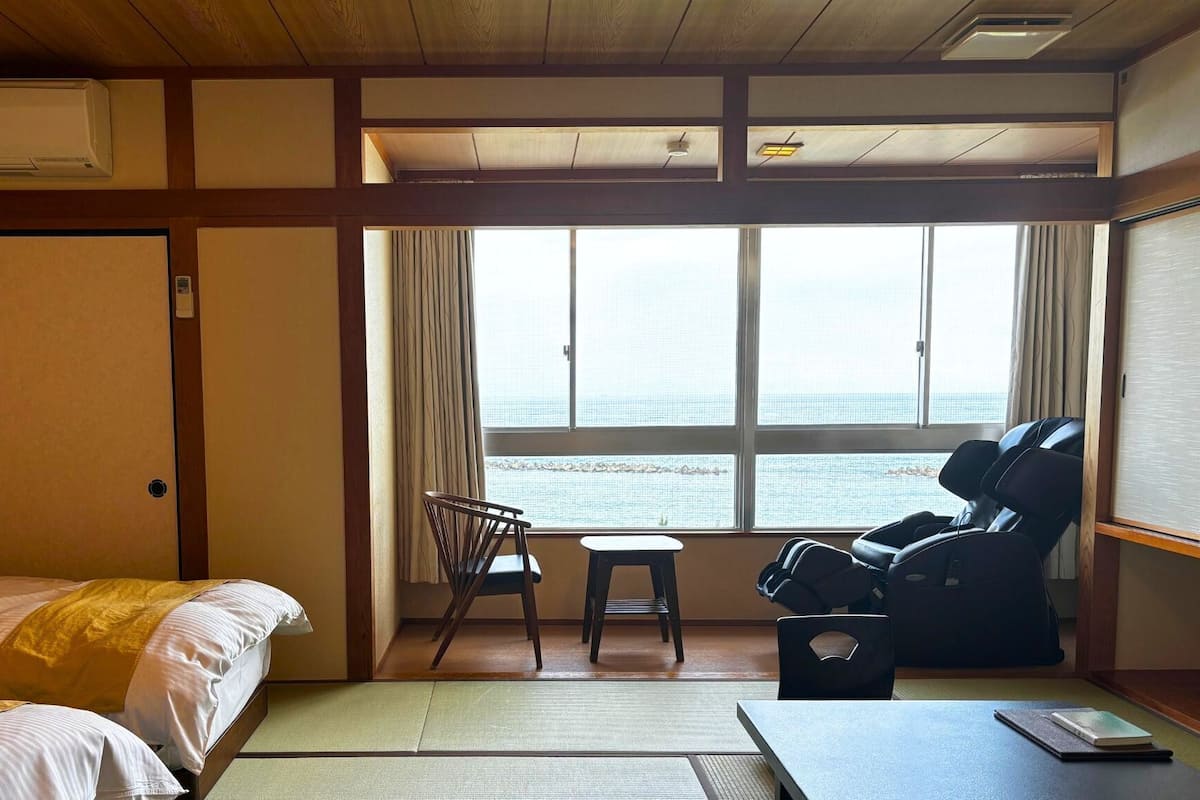 Habitación de hotel con cama, una silla, sillón de masajes, una mesita y vista al mar a través de amplias ventanas.