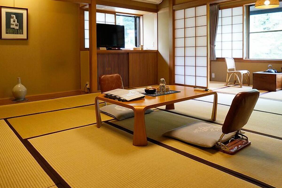 Um quarto japonês tradicional com piso de tatame, uma mesa baixa com almofadas, uma televisão e portas deslizantes shoji.