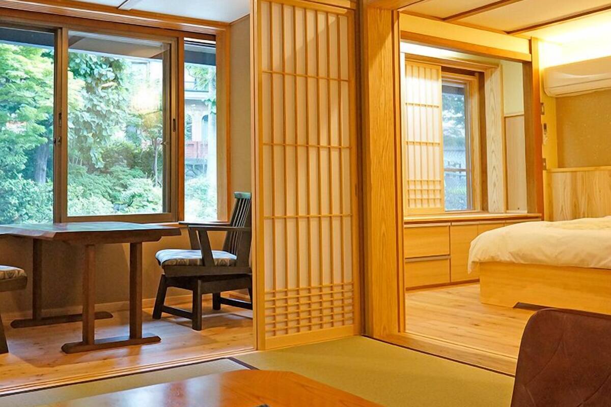 Um quarto tradicional em estilo japonês, com tatames, uma mesa baixa com uma cadeira, uma cama com cabeceira de madeira e portas corrediças com biombos shoji.