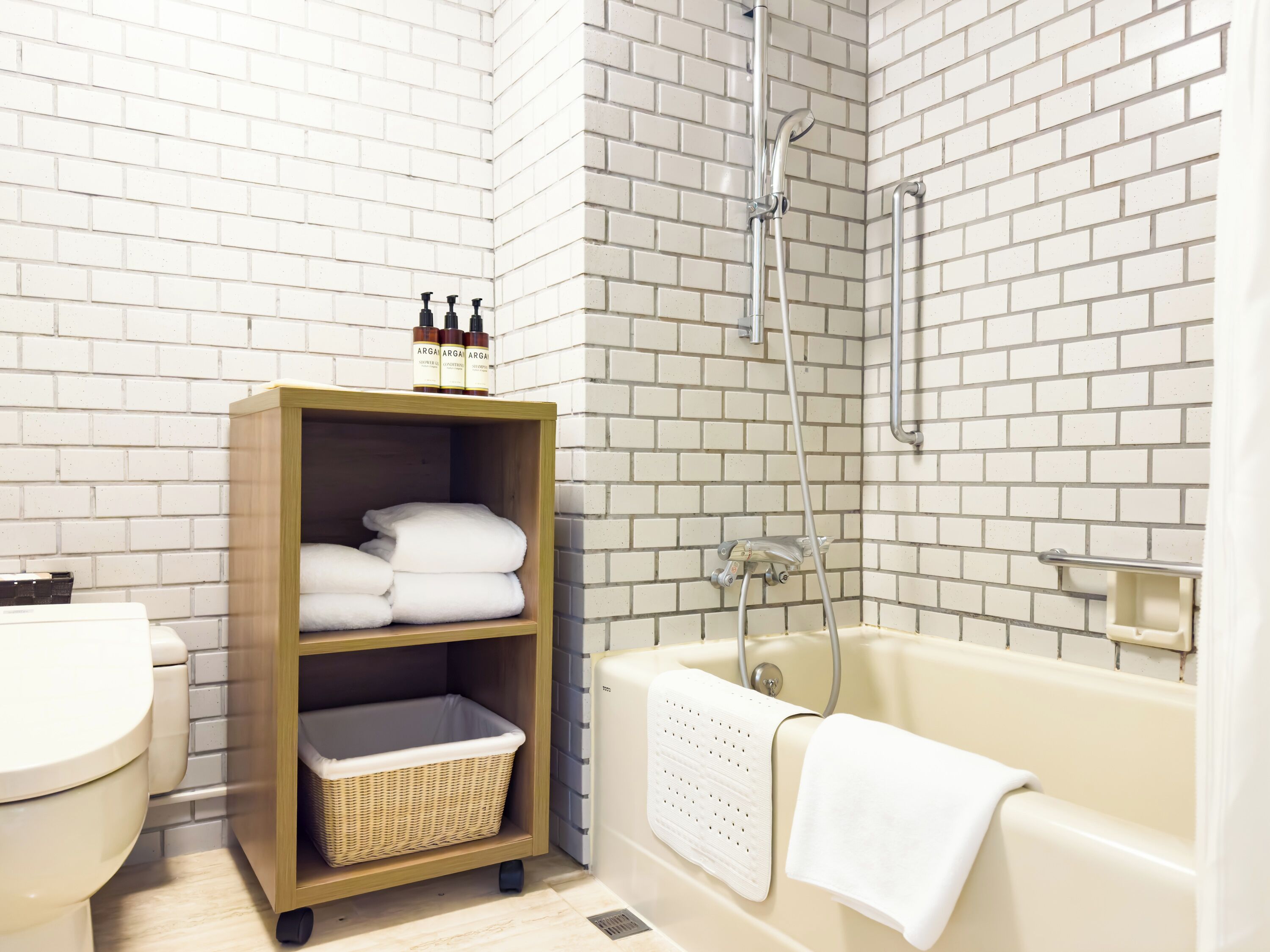 Une salle de bain avec un mur en briques blanches, une étagère en bois où sont rangés les articles de toilette, une baignoire blanche et des toilettes.