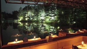 Un espacio de comedor al aire libre con bancos y mesas de madera, cada una con una vela, con vistas a un cuerpo de agua tranquilo y una zona boscosa.