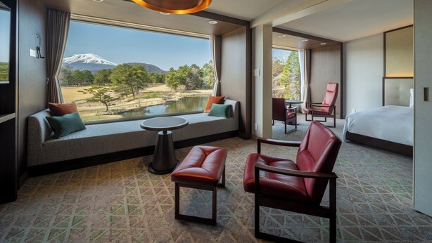 Hotelkamer met een groot raam, uitzicht op een berg en meer, een bed, een zithoek met een tafel en een rode lederen fauteuil.