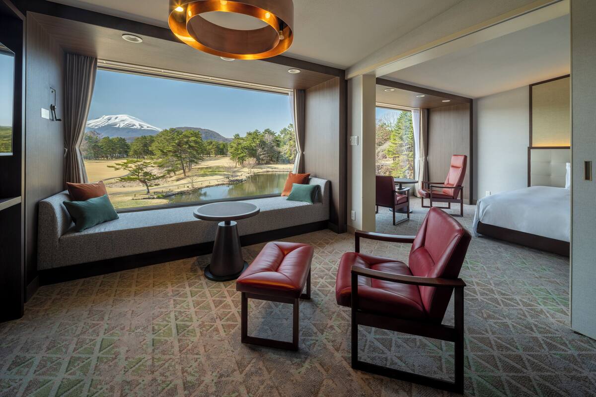 Camera d'albergo con una grande finestra che offre vista su una montagna e un lago, un letto, una zona salotto con un tavolo e una poltrona in pelle rossa.
