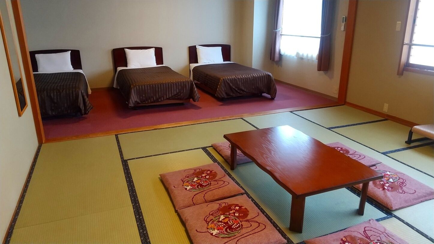 Quarto japonês tradicional com tatames, duas camas baixas, uma mesa de madeira e um banco.