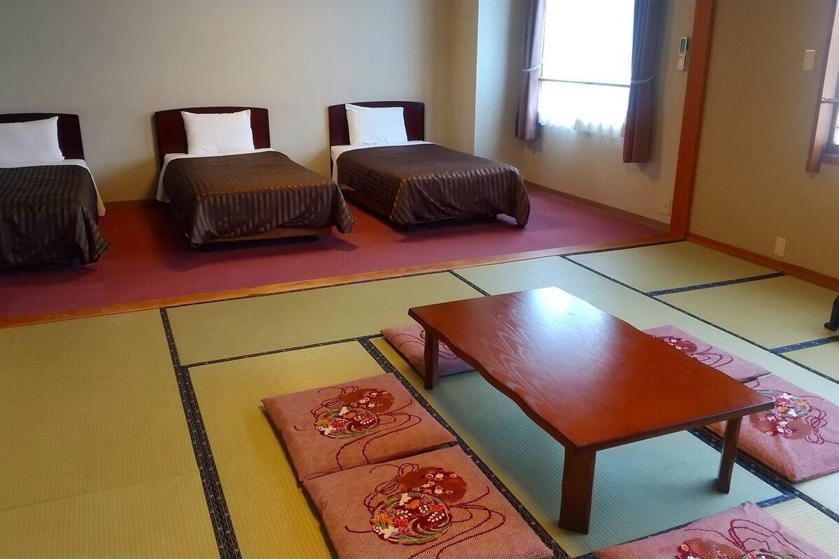 Una habitación japonesa tradicional con suelo de tatami, dos camas bajas, una mesa de madera y un banco.