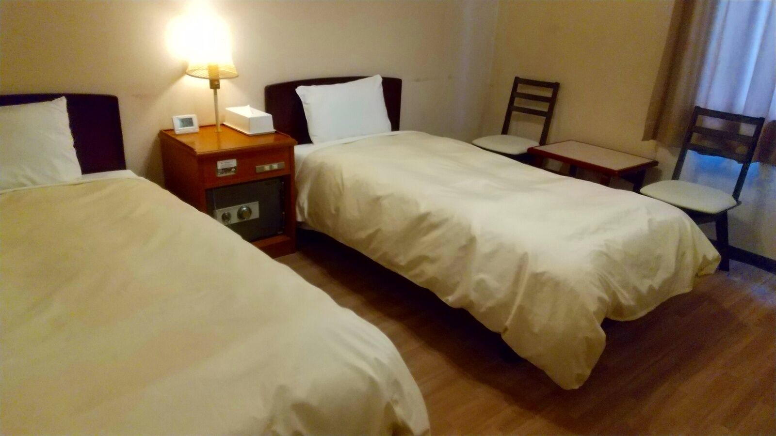 Habitación de hotel con dos camas, una mesita de noche con lámpara, una silla y una pequeña mesa.