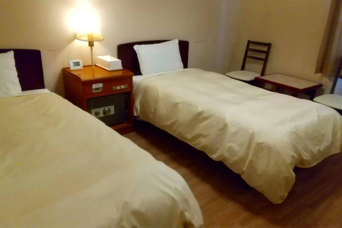 Habitación de hotel con dos camas, una mesita de noche con lámpara, una silla y una pequeña mesa.