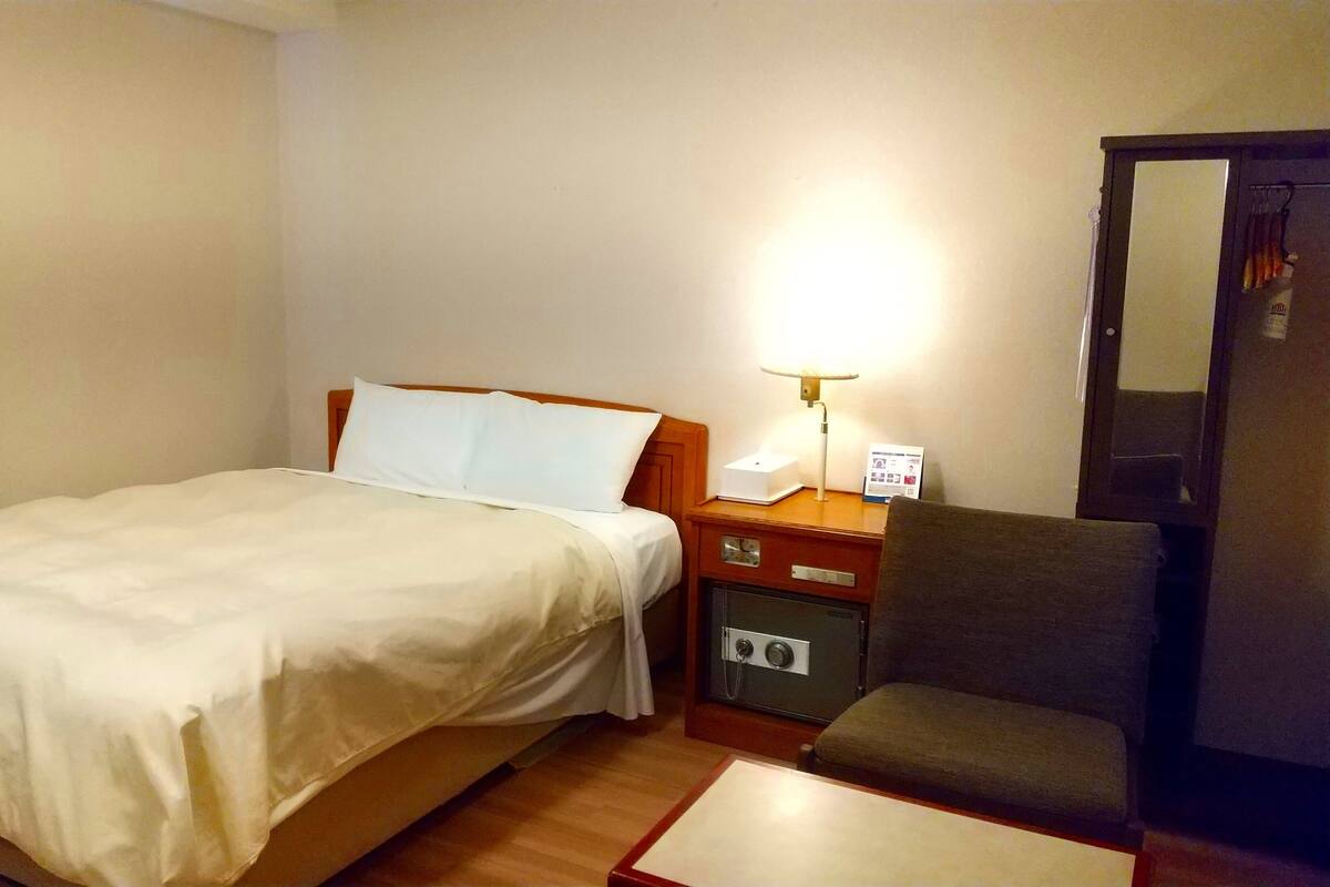 Una habitación de hotel con cama, escritorio con lámpara, una silla y armario.